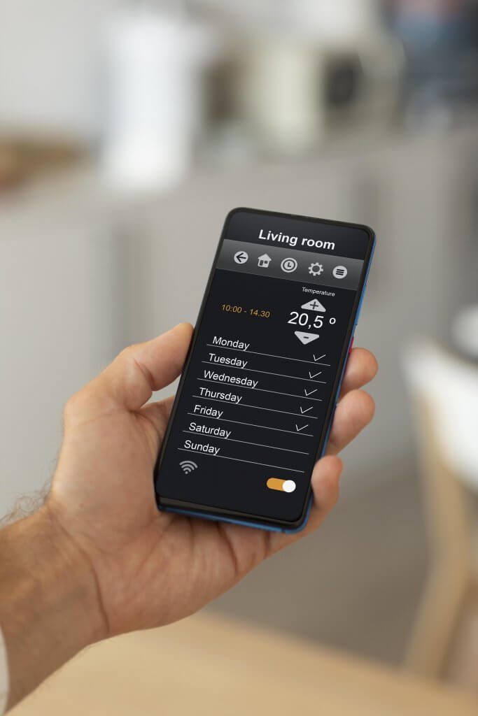 Eine Hand hält ein Smartphone mit geöffneter Kleiber GmbH App für Wohnraumlüftung, die Wochenpläne fürs Wohnzimmer zeigt