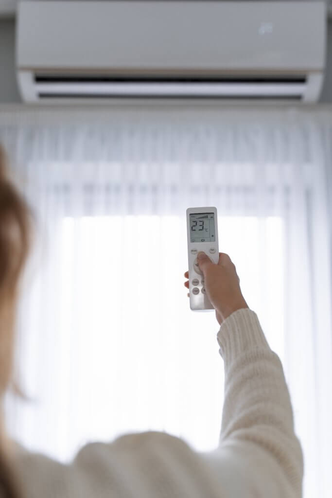 Eine Person stellt im sonnigen Karlsruher Einfamilienhaus mit einer Fernbedienung die Klimaanlage auf 23°C, Licht fällt durch Gardinen