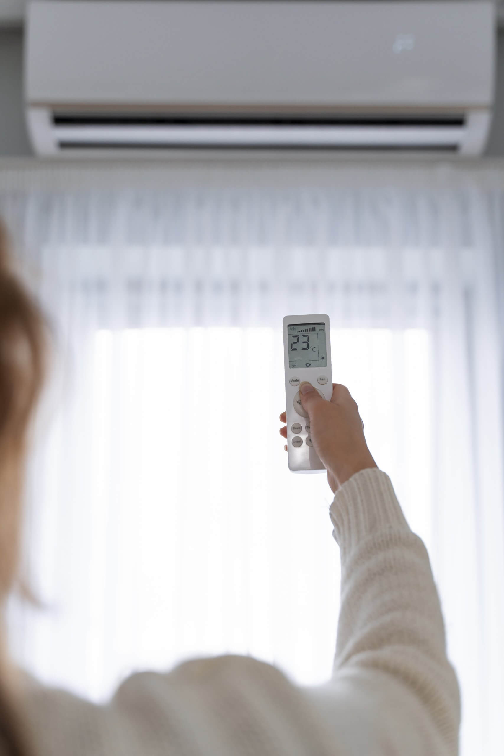 Eine Person stellt im sonnigen Karlsruher Einfamilienhaus mit einer Fernbedienung die Klimaanlage auf 23°C, Licht fällt durch Gardinen